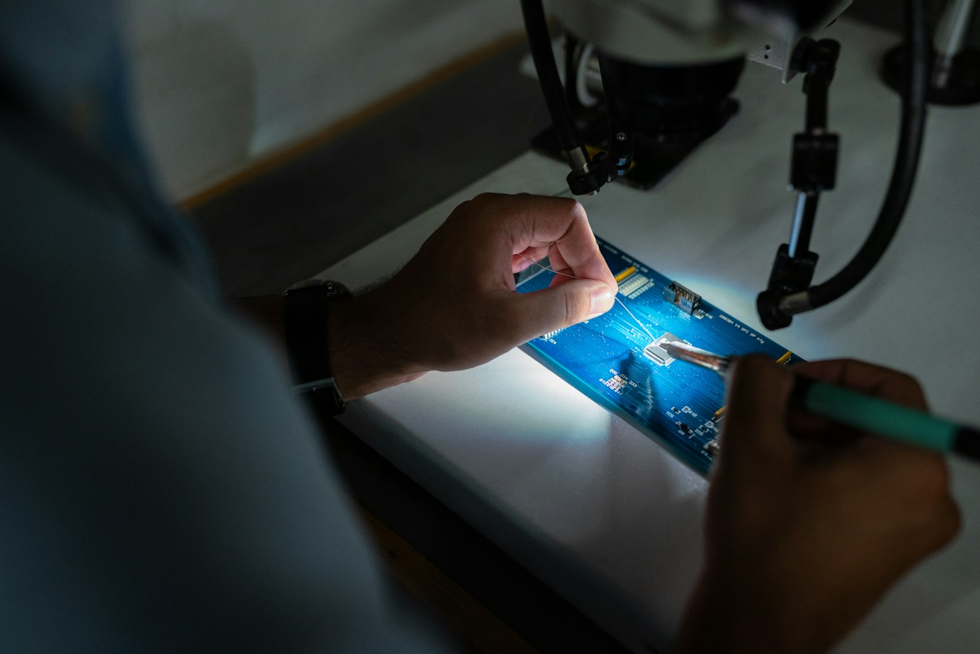 Hands working on an electronic circuit board beneath inspection equipment
